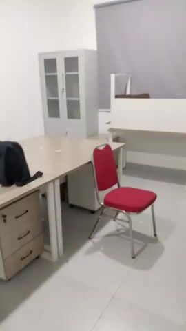 The video shows an empty office space. The room is lit and appears to be well-maintained. There are several office desks, some with drawers. One desk has a red chair in front of it. There is also a tall, grey cabinet with glass doors. The walls are white and the floor is tiled. A grey roller blind covers a window. In one shot, a black bag is seen on one of the desks.