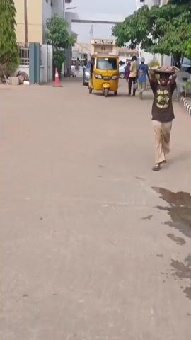 The video shows a scene in what appears to be an urban environment. A person is walking towards the camera, carrying a large tray or basket of goods on their head. The person is wearing a dark t-shirt and light-colored pants. In the background, there is a yellow auto rickshaw or tricycle vehicle, and other people are walking in different directions. The surroundings include buildings, trees, and a paved road. There is also what appears to be a traffic cone and a metal gate on the left side of the frame. The pavement has a few dark wet spots.