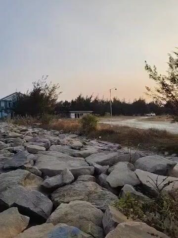 The video shows a rocky path leading towards a few buildings in the distance. The path is made of large, grey rocks of various sizes. On the left side of the path, there is a two-story building with a white facade and a blue roof. Behind the path, there are several trees and some smaller buildings. The sky is clear and a pale orange, suggesting it might be sunrise or sunset. A street light is visible on the right side of the path. The environment appears to be a coastal area, possibly near a beach, given the presence of rocks and the flat landscape.