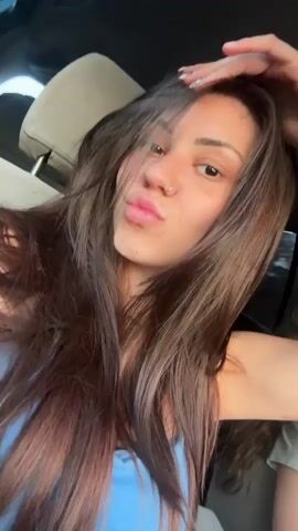 The video shows a young woman sitting in a car. She has long brown hair and is wearing a blue top. She looks at the camera, poses, closes her eyes, and winks. The interior of the car is visible in the background. The headrest of the driver's seat is visible, as well as the car window and frame.