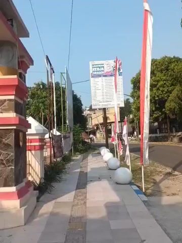 The video shows a sidewalk in front of a building with red and white decorative elements. Several Indonesian flags are displayed along the sidewalk. A large sign is mounted on a pole, with text that appears to be a list of names. There are also several spherical white bollards lining the sidewalk. Beyond the sidewalk, there is a road with some trees and buildings in the background. The sky is clear and blue. A motorcycle is visible on the road in the distance.