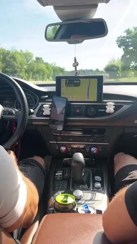 The video shows the interior of a car from the passenger's perspective, looking forward at the dashboard and the road ahead. A person's shoulder and arm are visible on the left, with their hand resting on the steering wheel. The steering wheel has an Audi logo. The car is in motion, as indicated by the blurred scenery outside and the speed displayed on the dashboard, which reads 135. On the dashboard, there is a large infotainment screen displaying a navigation map. A smartphone is mounted on the dashboard, also showing a navigation app. There are two small white teddy bear figurines on either side of the infotainment screen. A green can and a blue can are visible in the cup holders. The sky is blue with some clouds and contrails. The environment outside appears to be a road surrounded by trees and greenery.