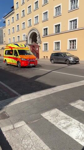 The video shows a yellow and red ambulance turning off of a road and onto a side street. The ambulance has the text "NUJNA MEDICINSKA POMOC" and the number "112" printed on its side. It also has a star of life emblem on the side. The ambulance is passing a zebra crossing. A gray SUV passes in front of the camera in the opposite direction of the ambulance. There is a pedestrian walking on the zebra crossing behind the ambulance. In the background, there is a building that says "DOVOZ URGENC." The sky is blue with white clouds. The ambulance has an EU license plate.