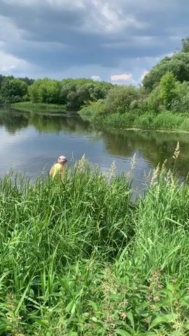 A person wearing a yellow shirt and a light-colored cap is standing in the shallow water of a river, partially obscured by tall green grass. They are holding a long fishing rod, which extends out over the water. The river is calm, with reflections of the green trees and bushes that line the banks visible on its surface. The sky above is partly cloudy, with patches of blue sky peeking through. The overall scene depicts a person fishing in a natural, serene environment.
