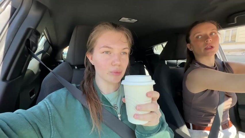 A woman in the passenger seat of a car drinks from a white paper coffee cup while wearing a green sweatshirt. She has brown hair pulled back into a ponytail. She is also wearing a silver necklace. The driver, a woman with brown hair in a ponytail, is wearing a brown tank top and white jeans. The car is in motion and passing buildings.
