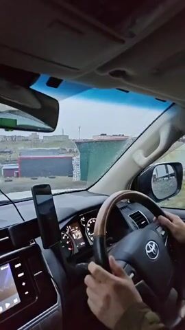 The video provides an interior view from a vehicle, looking forward through the windshield. The dashboard is visible, with a smartphone mounted to the left of the steering wheel. The steering wheel itself has wood accents and a Toyota logo. A driver's hands are visible on the steering wheel. A dashcam is mounted on the windshield, displaying "12." In the background, a somewhat industrial or rural landscape is visible, with various buildings, some appearing to be storage containers or sheds, and a dirt road. The sky is overcast. The vehicle is moving, as indicated by the blurring of the background and the movement of the steering wheel. The audio is muffled and appears to be a conversation.
