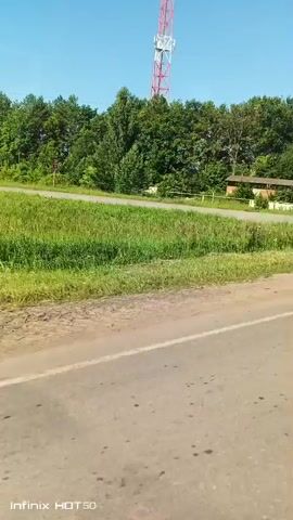 The video shows a blue station wagon driving on a dirt road next to a paved road. The car is blue and has a roof rack. The driver is visible inside the car. The environment is rural, with trees and a cell tower in the background. The weather appears to be sunny. The video is shot from a moving vehicle.