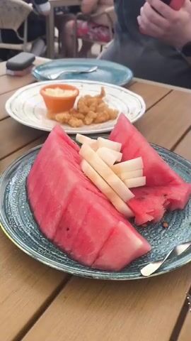 This video captures a meal being served at an outdoor restaurant, likely during the daytime given the natural light. The setting appears to be a wooden table in what could be a courtyard or patio area. In the foreground, a plate of sliced watermelon and white fruit, possibly pear, is presented with a fork. Further back, another plate holds two kebabs on skewers, served with flatbread, a small bowl of red sauce, and thinly sliced red onions. A separate plate in the background contains fried shrimp and a dip. There's also a glass of water on the table.

Two individuals are partially visible in the background, both appearing to be engaged with their phones. Their clothing suggests a casual atmosphere. The overall mood seems relaxed and convivial, typical of a leisurely meal.

The visible text on the metal skewers reads "HPERK". The video pans across the table, showcasing different dishes and the setting. The presence of watermelon, kebabs, and fried items suggests a diverse menu, possibly catering to various tastes. The Vahdat, Tajikistan location context implies a Central Asian culinary influence, which aligns with the presentation of the kebabs and flatbread. No specific time of day or weather conditions are explicitly discernible beyond the natural light.