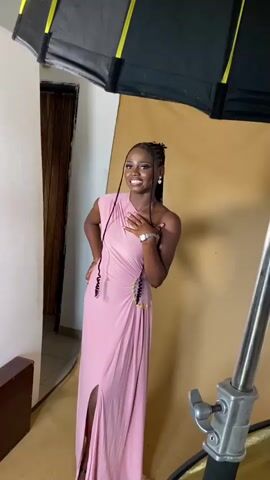 This media file captures a series of moments likely related to a photoshoot or video recording.

The first segment features a young Black woman posing in a pink, one-shoulder, floor-length gown with a high slit. She has dark braided hair, wears earrings and a watch, and has a bright smile, indicating a positive and celebratory mood. She is positioned in front of a warm, tan backdrop, with a large softbox light visible above her, suggesting professional lighting is being used.

The second segment shifts focus to a man wearing a black t-shirt and light-colored pants, engrossed in his phone while standing in what appears to be a living room. Behind him, an orange sofa and a camera on a tripod are visible, along with framed pictures on the wall.

The third and fourth segments zoom in on a young Black man wearing a blue Arsenal football jersey with "Emirates Fly Better" printed on it. He is looking off-camera with a thoughtful or perhaps slightly concerned expression. The same framed pictures seen in the previous segment are visible behind him, including what appears to be a wedding photo.

The overall setting appears to be indoors, possibly in a home or a small studio space in Anguldi, Nigeria. The lighting suggests the recording is taking place during the day. The activities suggest preparation for an event or a creative project.