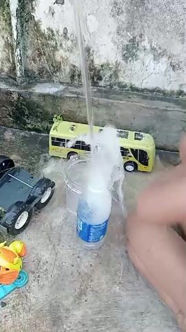 A close-up, low-angle shot captures a person's bare legs and feet on a concrete surface as water streams from above into a plastic bottle. The water is creating a foamy spray around the bottle's opening and overflowing into a clear plastic container. To the left of the bottle, a green toy bus and a gray toy truck are visible on the concrete. In the lower left corner, some colorful toy pieces are scattered on the ground. In the background, a textured wall or staircase rises. The scene suggests an outdoor setting, possibly a yard or patio.