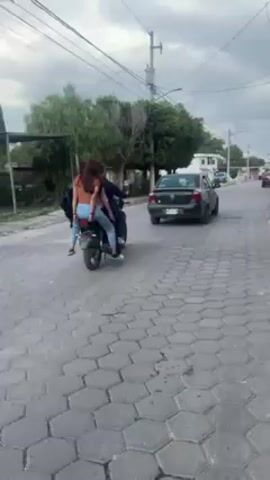 A motorcycle with three people on it is traveling down a street. The motorcycle is black, and one person in the back appears to be wearing a light blue top and jeans. A man who seems to be walking alongside the motorcycle wears a black top and dark pants. A black car is behind the motorcycle. The street is paved with hexagonal blocks, and there are buildings and trees along the side. The sky is overcast.