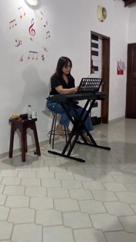 A woman sits at a keyboard in a tiled room with musical notes on the wall, playing a song and looking at sheet music. She is wearing a black shirt, jeans, and sandals. A stool is next to the keyboard stand with a bottle of water, bananas, and a microphone on it. A door with a piano keyboard painted on it is in the background. The tiles are white with a grid pattern.