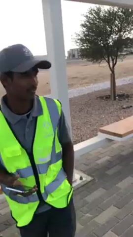The video shows three men in an outdoor setting. One man is wearing a grey baseball cap and a bright yellow-green safety vest over a grey polo shirt, and is holding a phone. He is standing near a white pillar structure and a wooden bench. Two other men are sitting on the bench. They are also wearing grey baseball caps and dark jackets. The ground is paved with grey bricks, and in the background, there is a gravel area and a bare tree. Further back, there is an undeveloped sandy landscape with a building visible in the distance. The sky is overcast. The man in the safety vest appears to be talking.