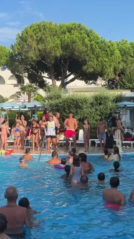 A group of people are gathered around a swimming pool on a sunny day. Several adults, including two men and two women, are dancing near the edge of the pool. One man is wearing red shorts and a bare chest, while the other is wearing a white t-shirt and black shorts. The women are wearing swimwear. Many children are in the pool, some with inflatable rings. Trees and buildings are visible in the background.