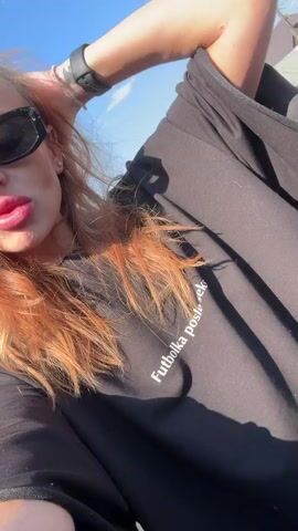 A young woman with long, reddish-orange hair and black sunglasses poses for a selfie video outdoors on a sunny day. She is wearing a black t-shirt with white text visible on the front, which reads "Futbolka posle seks". Throughout the video, she makes a pouting or "duck face" expression with her lips and occasionally runs her right hand through or on top of her hair. A clear blue sky with some clouds is visible in the background, along with a portion of a car's interior on the left side of the frame.
