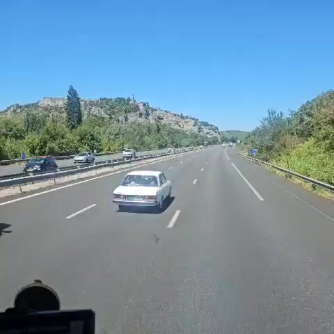 The video pans across a highway on a sunny day. In the foreground, a white sedan drives down the road. On the left side of the highway, a white truck is parked. In the background, a village with a church sits atop a hill. The sky is clear and blue.