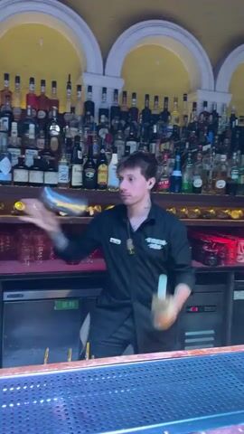 A bartender in a black shirt and apron juggles bottles behind a bar stocked with numerous bottles of liquor. The bar has a yellow wall with white arched accents and a metallic counter. The bartender's actions are blurred due to the rapid movement. Red lighting is visible behind the bar.
