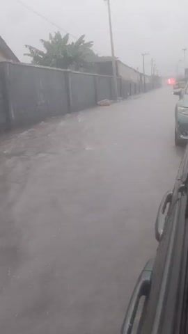 The video captures a street scene during what appears to be heavy rain or fog. The perspective is from inside a vehicle, looking out the side window and towards the road ahead. The road is wet and reflects the overcast sky. To the left, a long, dark wall runs parallel to the road, with some vegetation visible above it. Several utility poles are present along the side of the road. A car is parked on the right side of the road, with its hazard lights illuminated. Further down the road, another vehicle is visible, also with its lights on. The overall atmosphere is dim and hazy, suggesting poor visibility due to weather conditions.