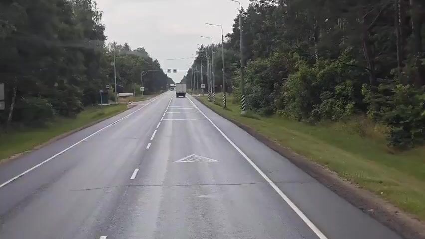 This video shows a view from a vehicle driving on a paved road through a wooded area. The road has multiple lanes, marked with white lines. There are trees lining both sides of the road, with some streetlights visible. In the distance, there is a truck driving in the same direction as the vehicle. The sky is overcast. The camera appears to be mounted on the dashboard or windshield of the vehicle, providing a forward-facing perspective.