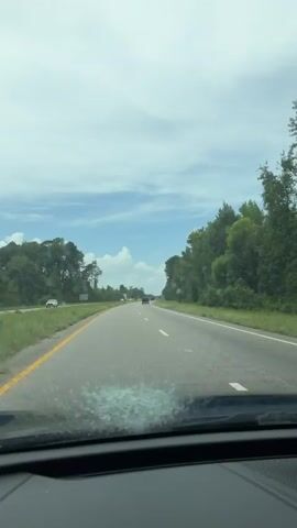 A first-person view from inside a vehicle shows a highway with trees lining both sides. The windshield is covered in what appears to be shattered glass and streaks of liquid, obscuring the view ahead. Windshield wipers are intermittently clearing small sections of the glass. Several other vehicles are visible on the road in the distance. The sky is cloudy.