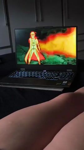 A laptop is open and placed on a dark surface, displaying an animated scene. The screen shows characters in a battle or intense situation. The laptop keyboard is illuminated with blue light. In the foreground, a person's legs are visible, suggesting they are sitting or lying down while watching the screen. The background appears to be a room with closet doors.