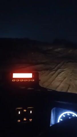 This is a first-person perspective video from inside a vehicle at night. The dashboard of the vehicle is visible, with illuminated gauges and a red display screen. The headlights of the vehicle are illuminating a rough, uneven terrain ahead, likely a dirt road or off-road path. The sky is dark with no visible stars or moon. The vehicle appears to be moving slowly through the landscape.
