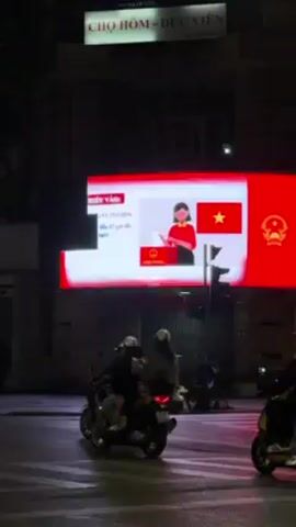 At night, a digital billboard with red and white content dominates the scene. In the background, a sign reads "CHỢ HÔM - ĐỨC VIÊN."  Several individuals on motorbikes are stopped at a traffic light, waiting to proceed.  A red car also drives through the intersection.  The overall environment is urban, with street lighting casting a glow on the scene.