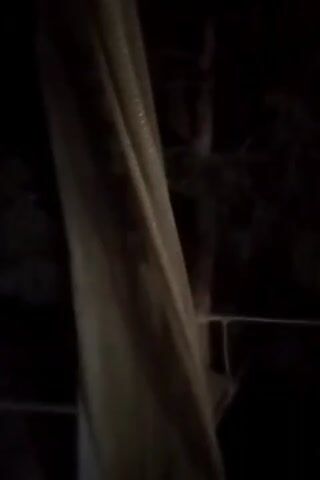 A close-up, low-light video shows a beige curtain on the left side of the frame. The curtain is hanging and appears to be indoors, possibly near a window. Through the gap in the curtain, a dark, blurry background is visible, suggesting trees and foliage outside. The lighting is dim, and no clear details of the environment or any individuals are discernible. The video captures a static scene, with no discernible movement or action.
