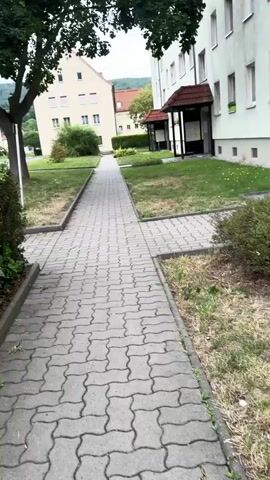 This is a video of a residential area. The camera is moving along a paved sidewalk that is lined with grass and trees. In the background, there are several apartment buildings. One building is a light beige color with a pitched roof, and another building is white with a red roof. There is also a street visible with cars parked along the side of it. The trees in the foreground are large and leafy, providing shade over the sidewalk. The sky is overcast, suggesting a cloudy day.