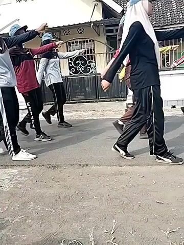 The video shows a group of people marching in formation on a paved street. The people are wearing uniforms, including hats and long pants. Some are wearing headscarves. They are marching in columns, with their arms extended forward. The background shows a house with a white wall and a tiled roof, and a fence. The lighting suggests it is daytime.