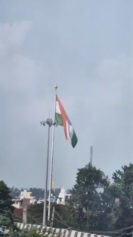 The video shows the Indian flag waving on a tall flagpole against a cloudy sky. A second, shorter flagpole with lights is visible to the right of the main flagpole. In the background, there are trees, buildings, and a telecommunications tower. A white car is parked on a road in the foreground. The camera appears to be moving slightly, possibly from a vehicle.