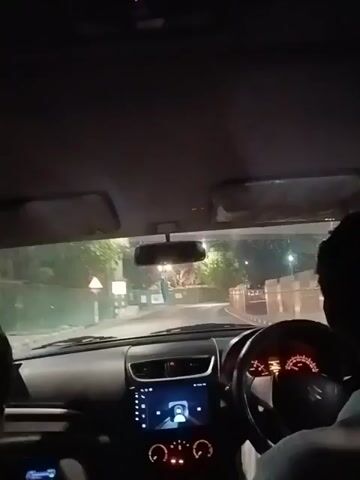 A driver is shown from behind, sitting in the driver's seat and driving a car at night. A passenger is partially visible in the front passenger seat. The car's dashboard, steering wheel, and speedometer are clearly visible. The car is driving along a street with trees and streetlights on either side. The video appears to have been shot from the back seat.