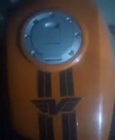 The video shows a close-up view of a motorcycle's fuel tank, which is orange with black stripes and a winged "V" emblem. The camera then pans up to the motorcycle's instrument cluster, featuring a speedometer, tachometer, and a digital display showing "0". The lights on the instrument panel are illuminated, indicating the motorcycle is ready to start. The scene appears to be outdoors at night, with dim lighting. The video ends with a shot of the illuminated instrument cluster against a dark background.