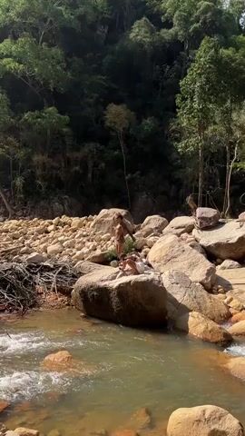 The video shows a scenic outdoor environment. The scene is dominated by a stream or river flowing amidst large rocks and boulders. Lush green trees and vegetation cover the surrounding hillsides. The camera pans up to reveal a tall waterfall cascading down a rocky cliff into a pool below. Several individuals are present in the scene, some standing on the rocks near the water and others swimming in the pool. The video captures the natural beauty of the location and the activities of people enjoying the surroundings.