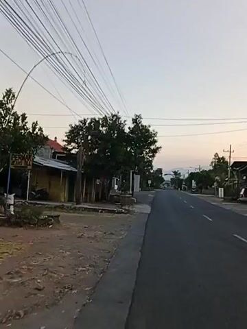 The video shows a paved road in a rural area with a clear sky and trees lining the sides. There are utility poles and wires running along the road. On the left side of the road, there is a building with a sign that reads "APOTEK DYAFARMA." The building appears to be a pharmacy or a shop. There is a sidewalk in front of the building. The road is empty, with no vehicles or people visible. The time of day appears to be early morning or late afternoon, as the light is soft and the sky is a pale orange.