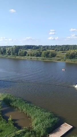 The video shows a scenic view of a wide river on a clear day with a blue sky and scattered clouds. In the foreground, there is a wooden dock surrounded by tall reeds. Further out in the river, a white motorboat with a blue canopy is cruising, leaving a wake behind it. There is also a smaller, lighter colored boat further away on the left side of the river, possibly a paddle boat or a small dinghy with people in it. The background features a lush green landscape with trees and some houses visible in the distance, suggesting a rural or semi-rural setting. The overall atmosphere is peaceful and idyllic.