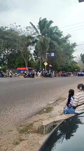 The video depicts a street scene with a crowd of people gathered on both sides of a road. On the left side of the road, several motorcycles are parked, and a group of people, some appearing to be law enforcement officers in uniform, are standing or milling about. On the right side of the road, closer to the camera, two people are seated on the curb, facing away from the camera. Further down the road on the right, a larger group of people are gathered, some of whom appear to be in uniform, possibly a marching band or a parade formation, holding flags. Palm trees and other vegetation are visible in the background, along with power lines and street signs. The sky is overcast. The overall atmosphere suggests an outdoor event or gathering.