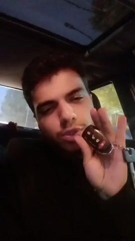 A man is sitting in the driver’s seat of a car, holding a car key fob near his mouth. The car is parked or stopped. The environment appears to be outdoors at dusk or night. The man is wearing a dark turtleneck shirt. He looks toward the camera.