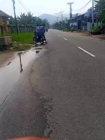 A motorcycle is parked on the side of a paved road. The motorcycle is dark in color and has a person wearing a blue shirt and dark pants sitting on it. The person appears to be looking down. To the left of the motorcycle, there is a patch of grass and a decorative fence. Further down the road, there are other motorcycles and a few buildings. The road is lined with power lines and poles. In the distance, there are mountains. The sky is overcast.