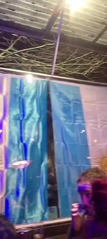 The video shows an outdoor gathering at night. In the foreground, there is a person's head and shoulders. In the background, there are two large flags hanging vertically, appearing to be the flag of Argentina due to the sun emblem on the left flag and the blue and white stripes. The area is lit by purple and blue lights, suggesting a festive or celebratory atmosphere. There are also structural elements visible overhead, possibly a roof or canopy, and some trees or branches. Further in the background, a group of people are gathered near what appears to be a building or establishment with colorful graffiti and signage. The lighting creates a vibrant, possibly club-like or party environment.