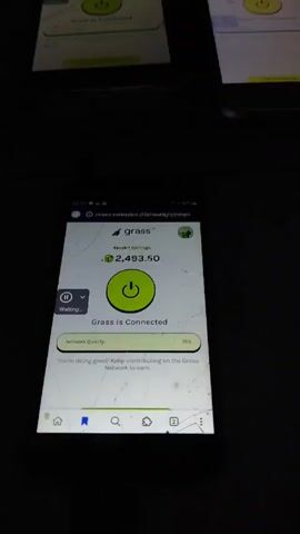 Okay, here's a forensic description of the video:

The video is shot in portrait mode, displaying the screen of a black smartphone. The smartphone screen is cracked in multiple places. The screen displays the "grass" app, showing an epoch earnings amount of 2,493.50. A large green power button icon in the center of the screen indicates "Grass is Connected." The network quality is reported as 75%. Text below states, "You're doing great! Keep contributing on the Grass Network to earn." There is a pause/waiting button on the top left of the screen. At the bottom is a navigation bar with home, bookmark, and a number 2 icon. The phone appears to be on a dark or black surface. 

Audibly, a strong wind can be heard.