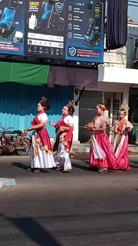 A group of children in traditional dance costumes are parading down a street. The costumes consist of white dresses with red sashes and gold headpieces. They are walking in a line, performing synchronized movements. Behind them, other people in traditional attire are also walking. The street is paved and bordered by buildings with shops and advertisements. The weather appears to be sunny.