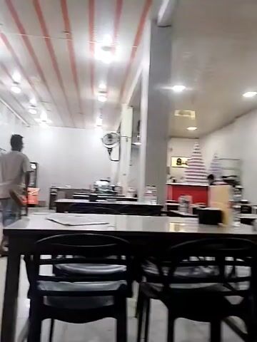 The video shows the interior of a restaurant. In the foreground, there are several tables with chairs. In the midground, a man is walking away from the camera. In the background, there is a counter with what appears to be drinks or bottles stacked in a pyramid shape. The ceiling has alternating orange and white stripes with lights. Fans are mounted on the walls. The overall impression is a clean and well-lit dining area.