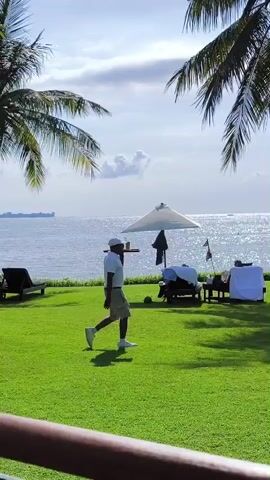 A person wearing light colored clothing, including a white hat, is walking across a green grassy area towards the sea. The sea is visible in the background, along with palm trees, lounge chairs and a white umbrella. The sky is blue with scattered clouds.