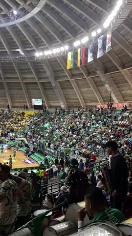 The video shows an indoor basketball arena filled with spectators. The stands are predominantly green and yellow, suggesting team colors. The court is visible with players in green uniforms practicing or warming up. Several national flags, including Venezuela, are hanging from the ceiling. The atmosphere appears to be of a live sporting event, with a large crowd engaged in watching the game. Some individuals in the foreground are wearing green shirts and appear to be part of the cheering section, with at least one person holding a drumstick. The lighting suggests it is daytime or an indoor event with artificial lighting. There are scoreboards and advertisements around the arena.