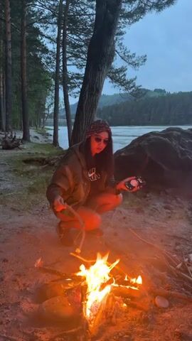 A woman with long dark hair, wearing sunglasses, a dark hooded jacket, and a patterned headband, crouches on sandy ground beside a small, brightly burning campfire. She holds a stick with a food item, possibly a sausage, over the flames with her right hand. In her left hand, she holds a black smartphone with a pink case. She looks towards the camera. Behind her, a large tree trunk stands, and a forest line is visible. A wide river flows in the background, with distant hills or mountains under an overcast, twilight sky.