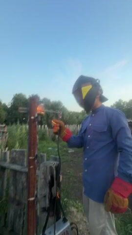 A person wearing a blue long-sleeved shirt, tan pants, a welding mask, and protective gloves is welding a rusty metal pole outdoors. Sparks are flying from the welding torch. A welding machine is visible at the bottom of the frame. The background shows a wooden fence, trees, and a clear blue sky. The scene appears to be in a rural or backyard setting.