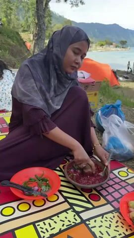 A young woman wearing a gray hijab and a dark purple dress is sitting on a colorful mat outdoors, next to a body of water with mountains in the background. She is using a mortar and pestle to mash red food, likely a condiment or dish, with her right hand. In front of her, on the mat, are two red plates. The plate to her left contains green chilies and sliced red onions. The plate to her right appears to have more food, possibly fruits or vegetables. To her right, there are plastic bags and an orange tent. In the background, there are other people, some near the water, and a person holding a child. The overall scene depicts an outdoor picnic or gathering by the lake.