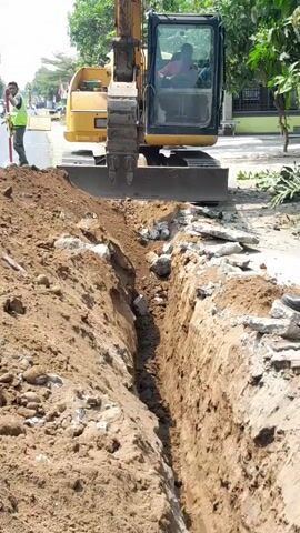 An excavator is digging a trench in the ground next to a paved road. A person wearing a high-visibility vest stands to the left of the trench, holding a long pole. Another person is visible in the cab of the excavator. Debris, including chunks of concrete, is scattered around the edge of the trench. Trees and buildings are visible in the background.