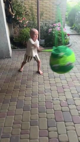 A young, bald boy with fair skin is playing outdoors in a courtyard. He is wearing a white t-shirt with a graphic design and striped shorts. He is barefoot and is swinging a stick at a large, round, green piñata. The piñata is decorated with lighter green swirls and appears to be made of paper. The boy is in the process of striking the piñata with the stick, which is also green. He has a determined expression on his face.

The setting is a paved courtyard made of interlocking rectangular stones in various shades of red, tan, and grey. Behind the boy, there is a brick wall and a thin metal pole. To the left of the boy, a portion of a building with a dark gray textured wall is visible. In the background, there are lush green plants and pink hydrangeas, suggesting a garden area. The lighting indicates it is daytime, likely sunny given the shadows. The overall atmosphere appears to be one of playful activity and outdoor recreation, possibly a birthday celebration. No visible text is present in the media file. The location is Dvorichnyi Kut, Ukraine.